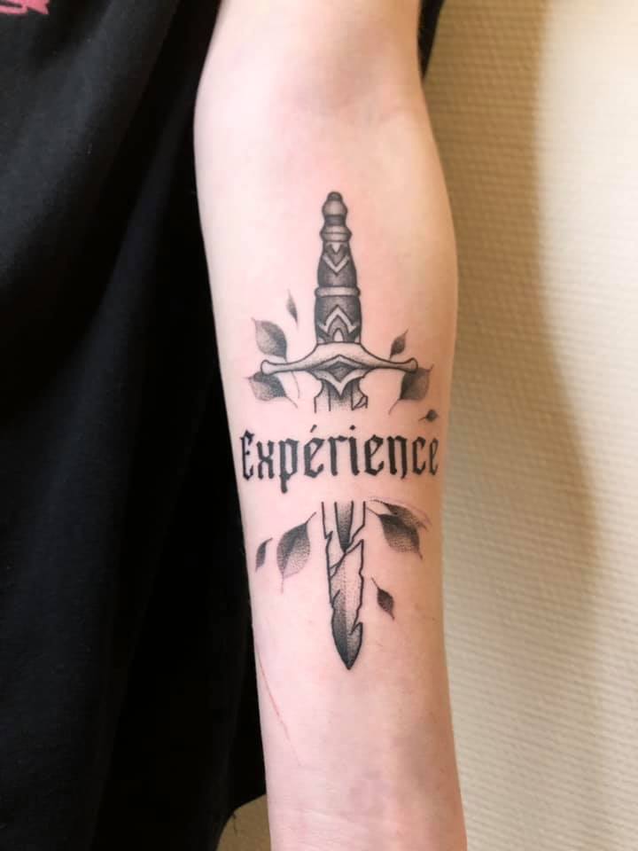 Un tatouage d'une épée en travail de points avec le mot "expérience" la traversant