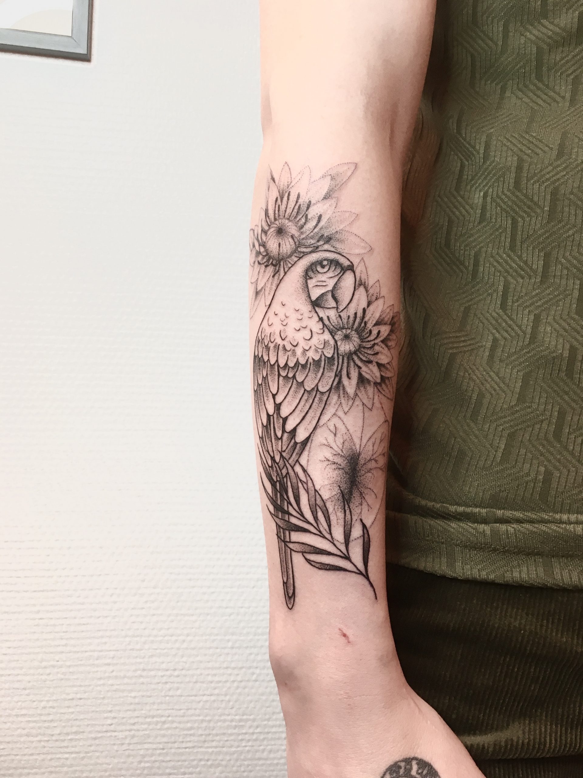 Un tatouage d'un perroquet et des fleurs sur l'avant-bras, le tout en travail de points